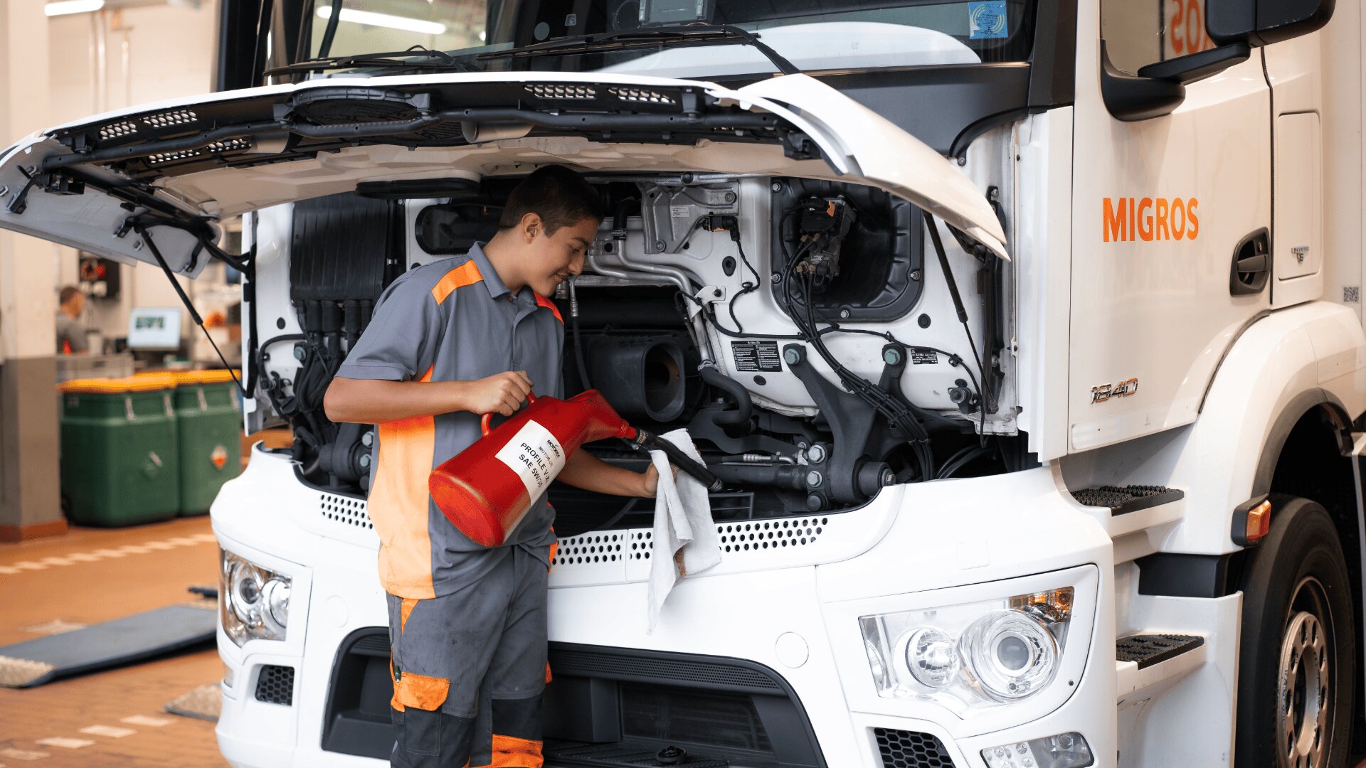Ein Mechaniker in grauer und orangefarbener Uniform arbeitet an einem weißen Lastwagen mit offener Motorhaube, der einen roten Ölbehälter und ein weißes Tuch in einer Werkstatt hält.