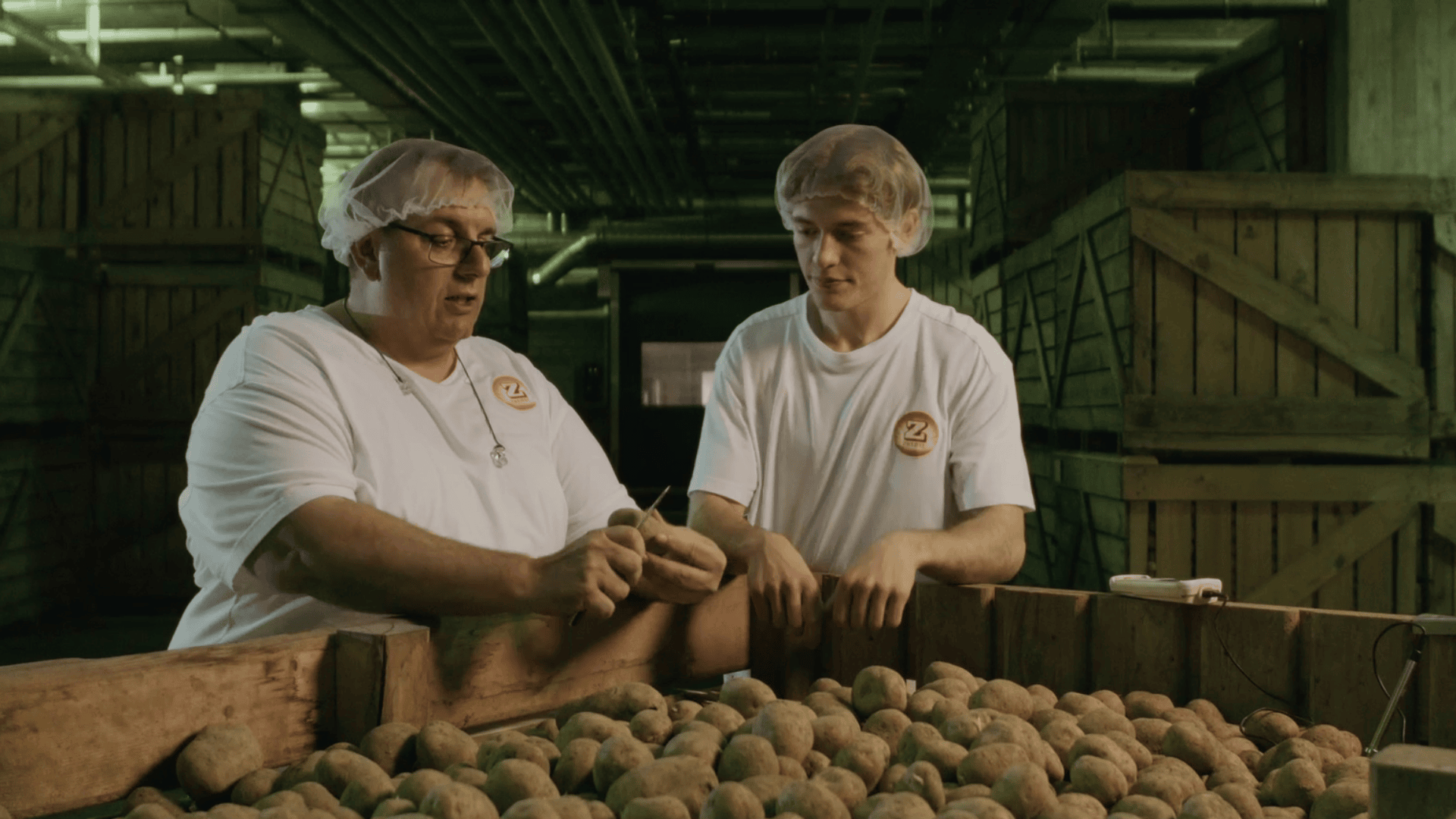 Zwei Arbeiter mit Haarnetzen und weißen Hemden sortieren Kartoffeln in einem Lagerhaus. Der eine hält eine Kartoffel, während der andere sie beobachtet, umgeben von Kisten, die mit weiteren Kartoffeln gefüllt sind. Die Umgebung ist industriell beleuchtet und hat grünliche Farbtöne.