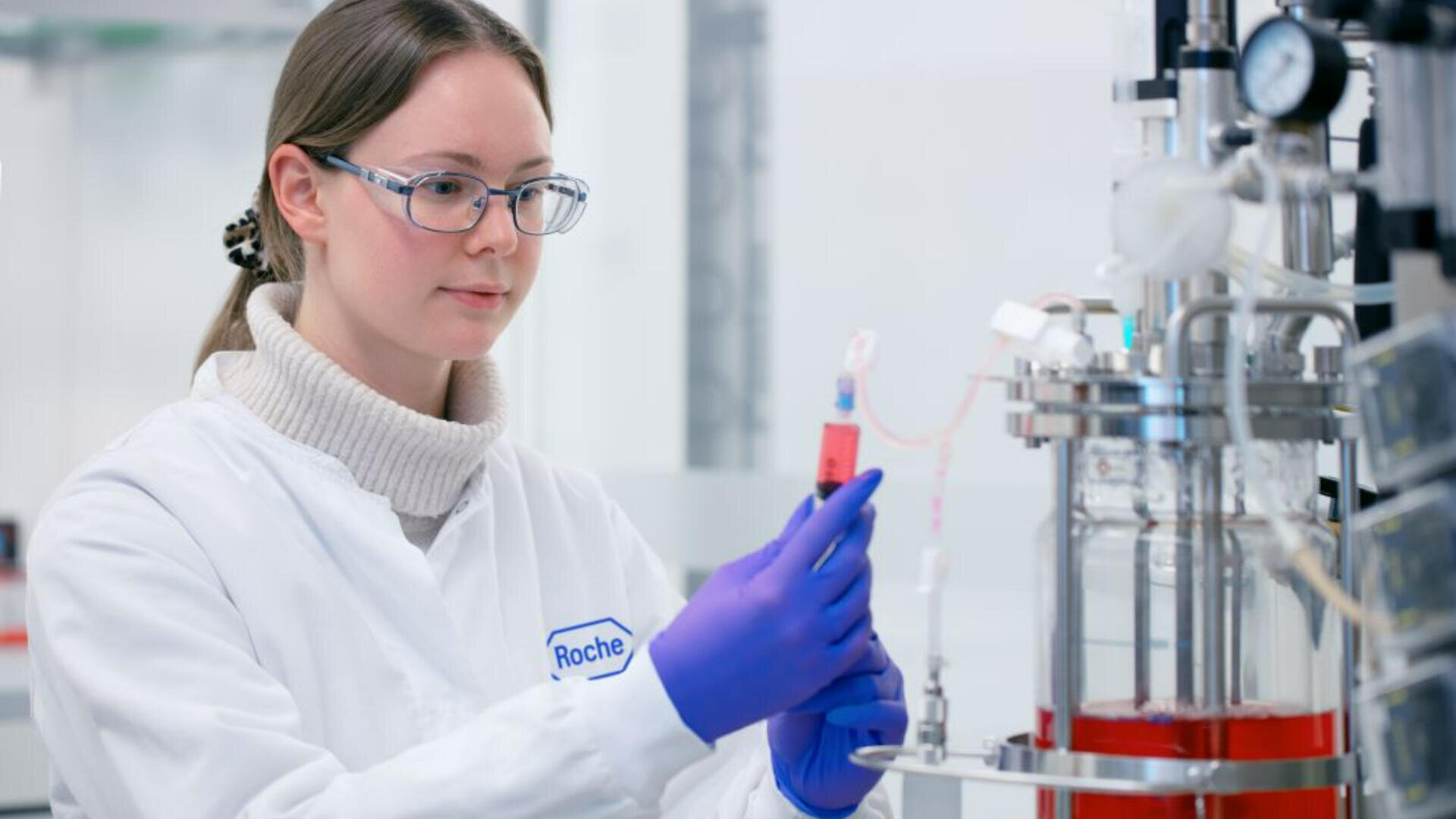 Ein Wissenschaftler, der eine Brille, Handschuhe und einen Laborkittel mit der Aufschrift Roche trägt, verwendet eine Spritze, um in einem sauberen, hellen Labor Flüssigkeit in die Laborgeräte zu füllen.
