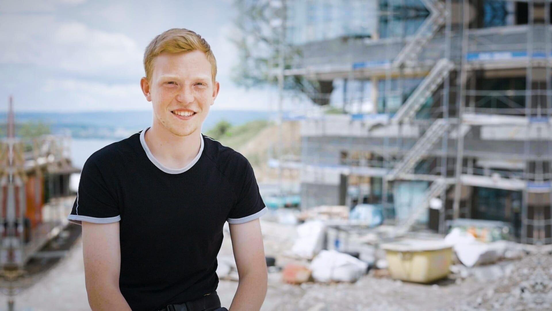 Ein junger Mann mit kurzen hellbraunen Haaren, der ein schwarzes T-Shirt mit hellem Saum trägt, lächelt, während er auf einer Baustelle mit Gerüsten und Baumaterialien im Hintergrund sitzt.