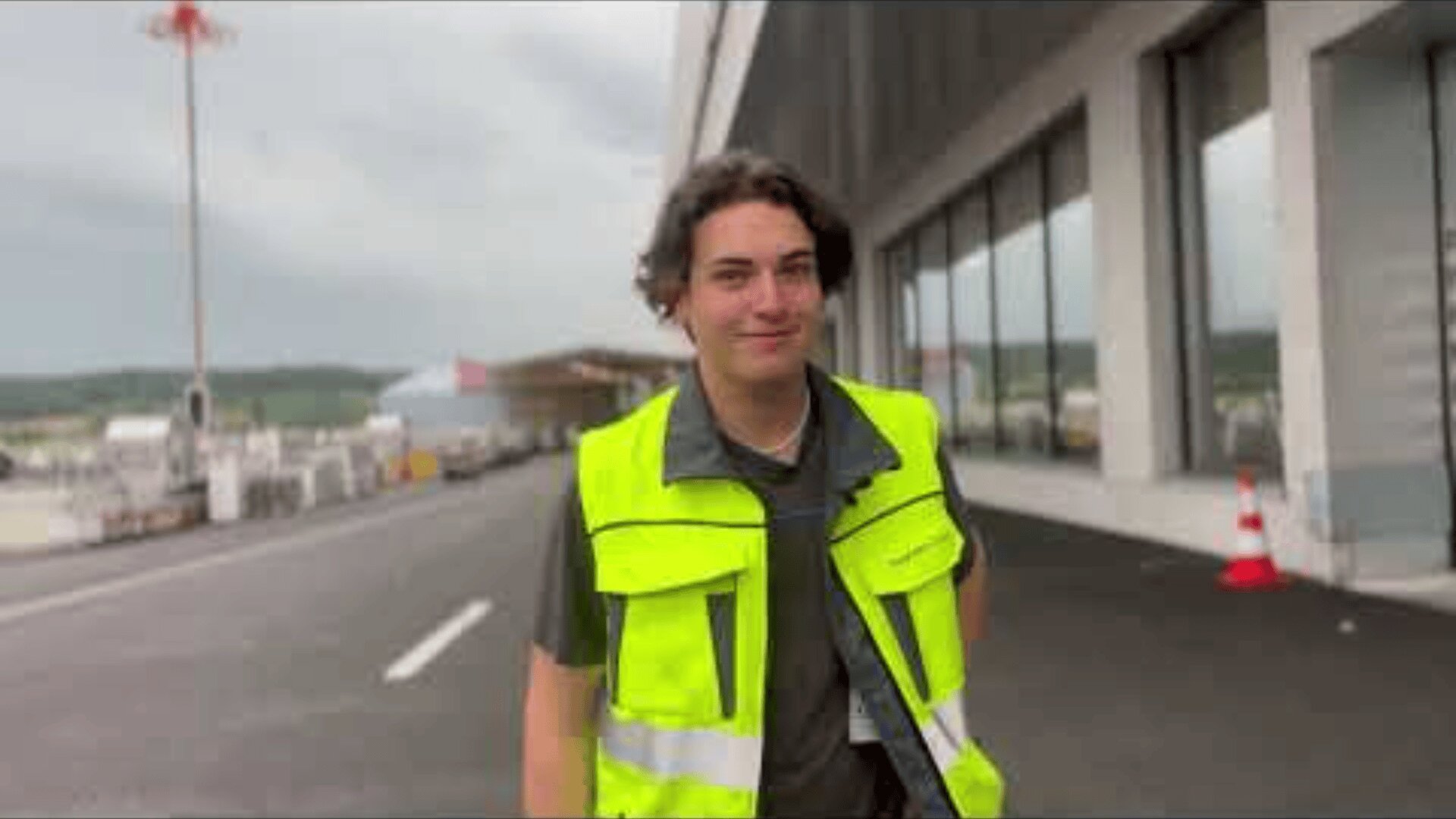Eine Person, die eine leuchtend gelbe Sicherheitsweste trägt, steht draußen in der Nähe eines Gebäudes, wahrscheinlich an einem Industrie- oder Flughafenstandort, wobei im Hintergrund Straßenlinien und Verkehrskegel zu sehen sind.