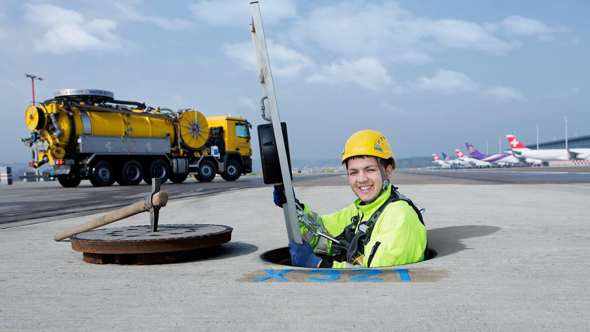 Ein lächelnder Arbeiter mit gelbem Helm und Warnweste kommt aus einem Gully auf dem Rollfeld eines Flughafens und hält den Deckel. Im Hintergrund sind ein großer gelber Servicewagen und Flugzeuge zu sehen.