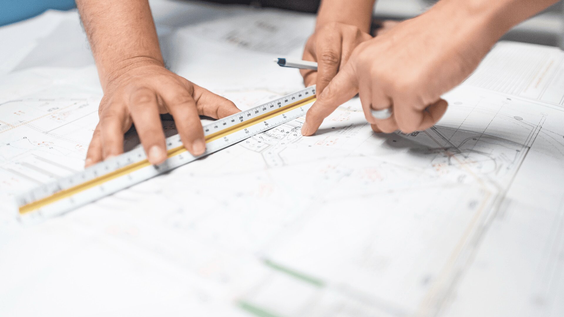 Zwei Hände arbeiten über Architekturplänen; eine Hand hält ein Lineal, während die andere mit einem Stift auf einen Ausschnitt der Pläne zeigt, was auf Zusammenarbeit und Entwurfsarbeit hindeutet.