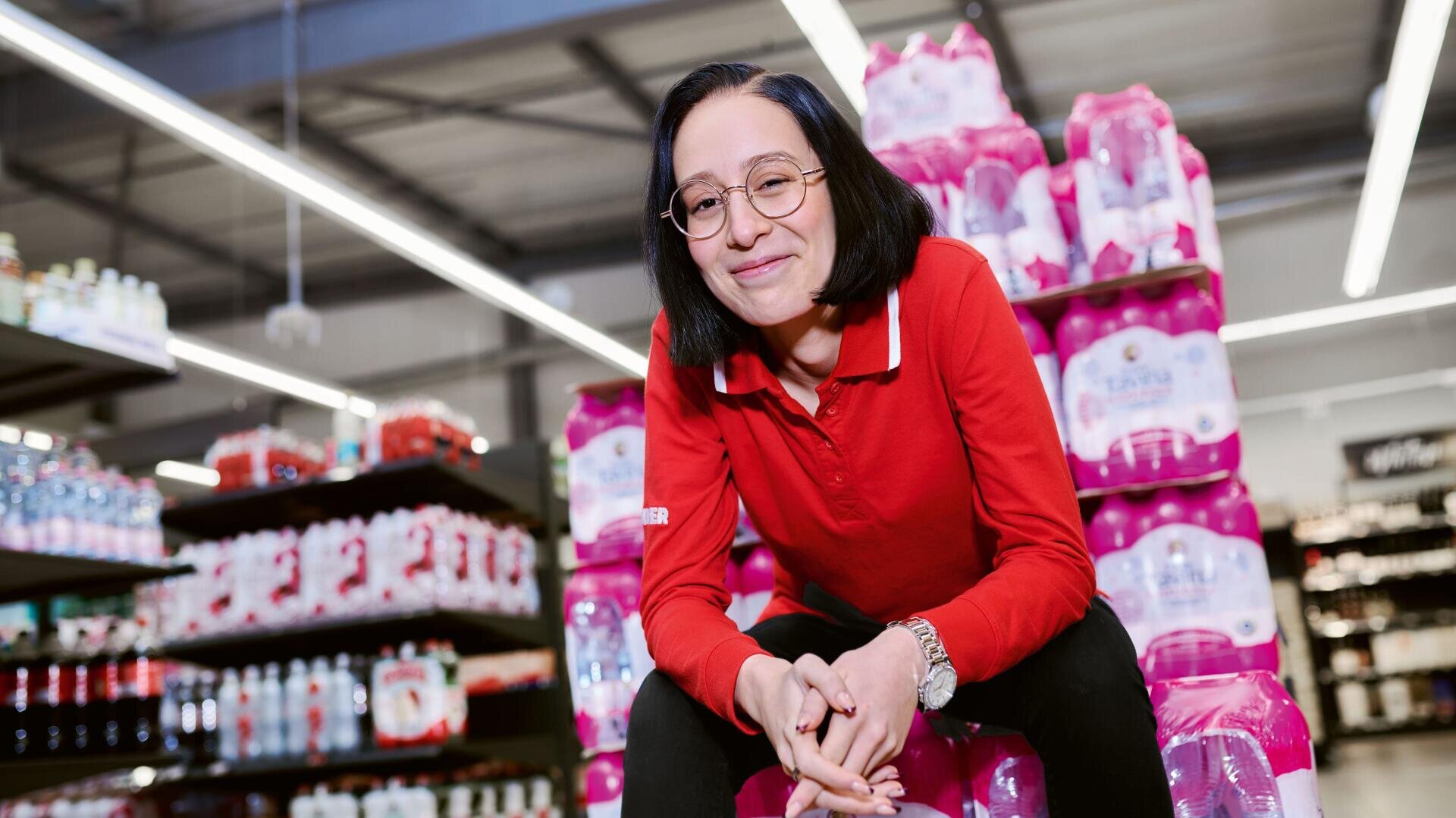 Eine Person mit Brille und rotem Hemd lächelt, während sie in einem hell erleuchteten Supermarktgang auf gestapelten rosa Wasserpackungen sitzt. Im Hintergrund sind Regale mit Getränken und Produkten zu sehen.