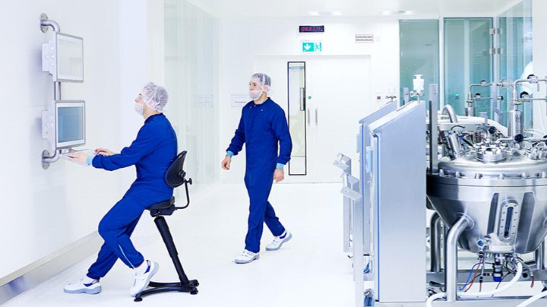 Zwei Personen in blauen Overalls und Haarnetzen arbeiten in einem sauberen, modernen Labor; eine bedient einen an der Wand montierten Computer, während die andere an Laborgeräten aus Edelstahl vorbeigeht.
