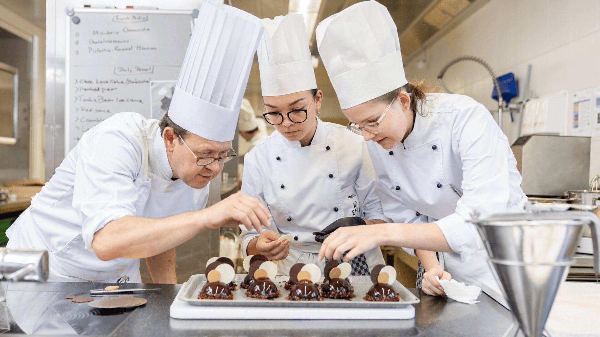 Drei Konditoren in weißen Uniformen und mit hohen Hüten dekorieren sorgfältig Schokoladendesserts auf einem Tablett in einer professionellen Küche, während im Hintergrund ein Whiteboard und Utensilien zu sehen sind.