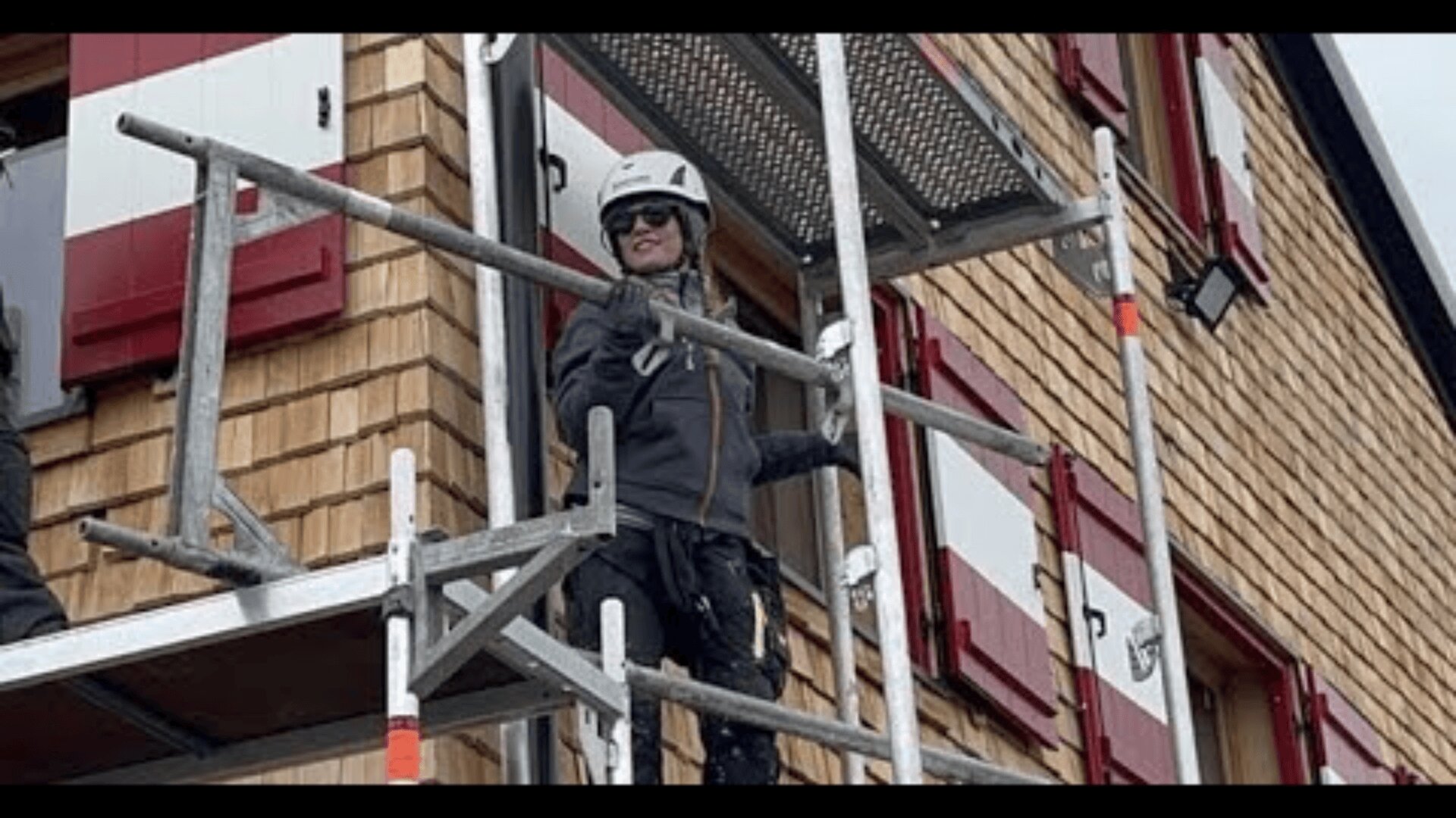 Eine Person, die einen Helm, eine Sonnenbrille und Arbeitskleidung trägt, steht auf einem Gerüst, das an der Seite eines Holzgebäudes mit roten und weißen Fensterläden befestigt ist.