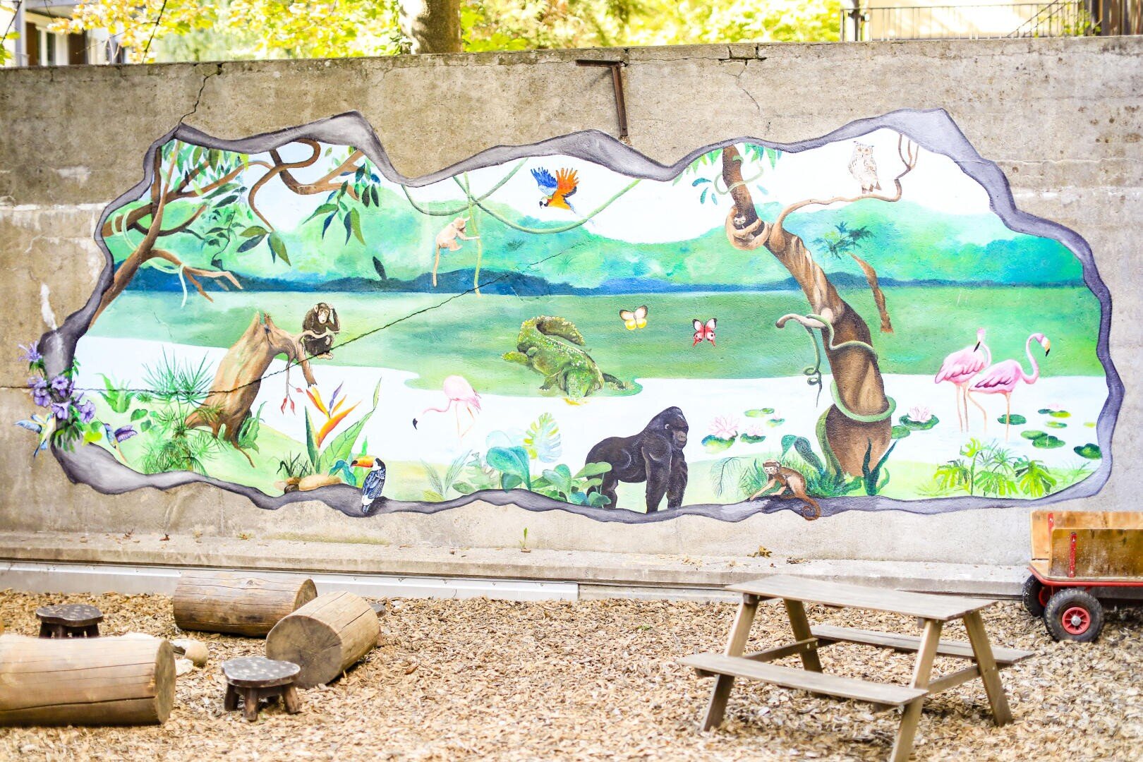 Ein auf eine Betonwand gemaltes Wandbild zeigt eine Dschungelszene mit Tieren wie einem Gorilla, Flamingos, Affen, Fröschen, Schmetterlingen und Vögeln, umgeben von einem Risseffekt; davor stehen ein Picknicktisch und Baumstämme.