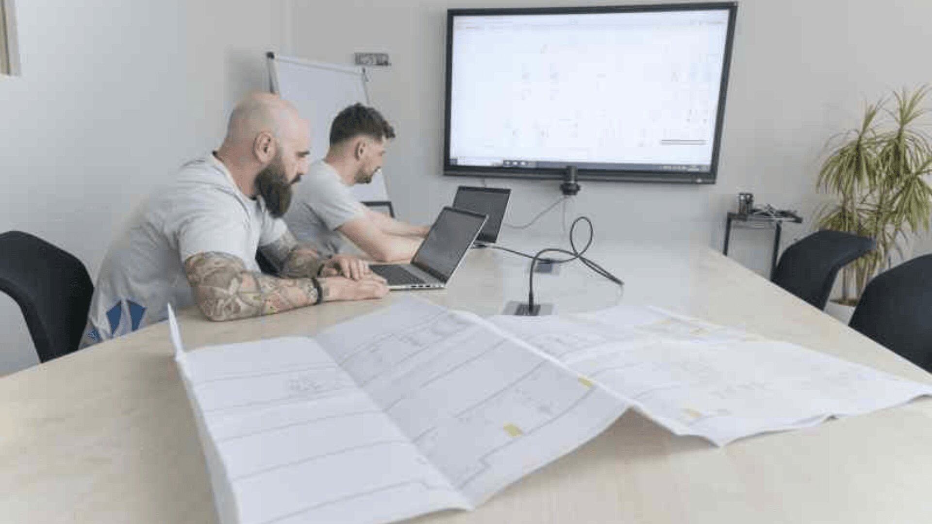 Zwei Männer arbeiten an Laptops an einem Konferenztisch, auf dem große Baupläne ausgebreitet sind. Auf einem Bildschirm an der Wand sind Diagramme zu sehen, und im Hintergrund stehen ein Whiteboard und eine Topfpflanze.