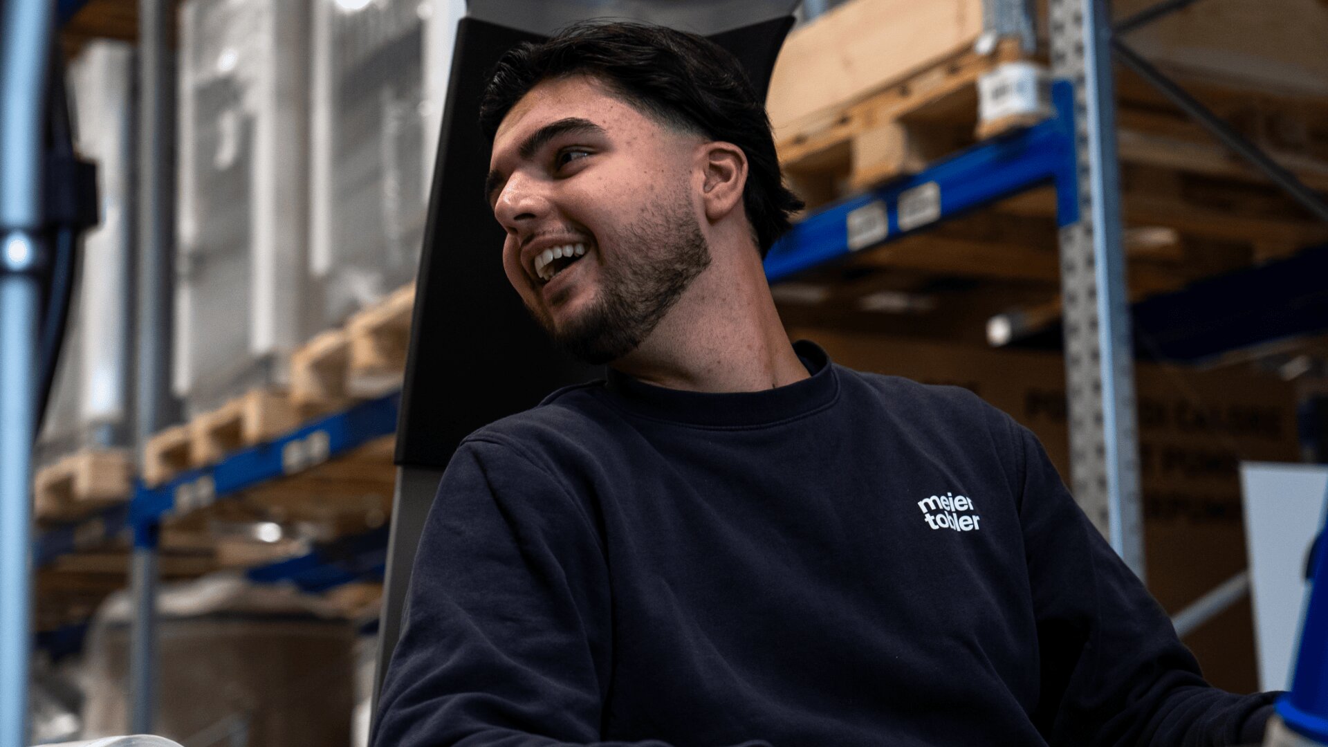Ein junger Mann mit dunklem Haar und Bart lächelt und blickt zur Seite, während er in einem Haus sitzt. Er trägt ein dunkles Sweatshirt mit weißer Schrift. Im Hintergrund sind Regale mit Kisten zu sehen.