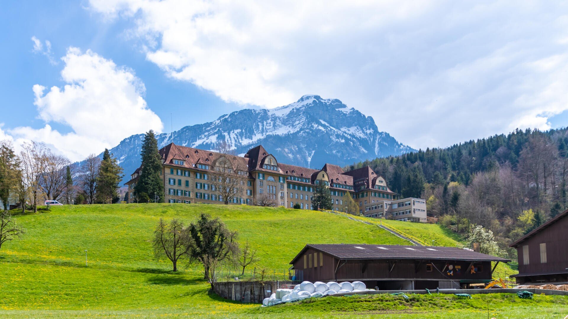 Großes historisches Gebäude mit rotem Dach auf einem grünen Hügel, hinter dem sich bewaldete Hänge und ein schneebedeckter Berg unter einem teilweise bewölkten Himmel befinden. Im Vordergrund sind eine Scheune und Ackerland zu sehen.