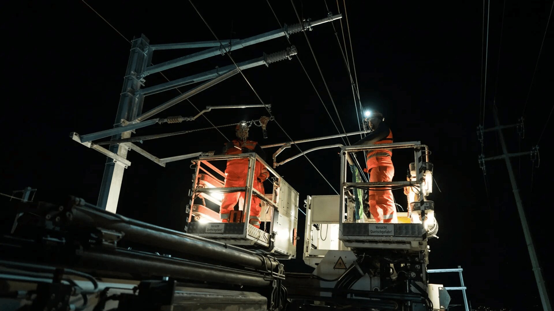 Zwei Arbeiter in orangefarbenen Overalls und Helmen stehen nachts auf einer erhöhten Plattform und reparieren Freileitungen, während helle Lichter ihren Arbeitsbereich beleuchten.