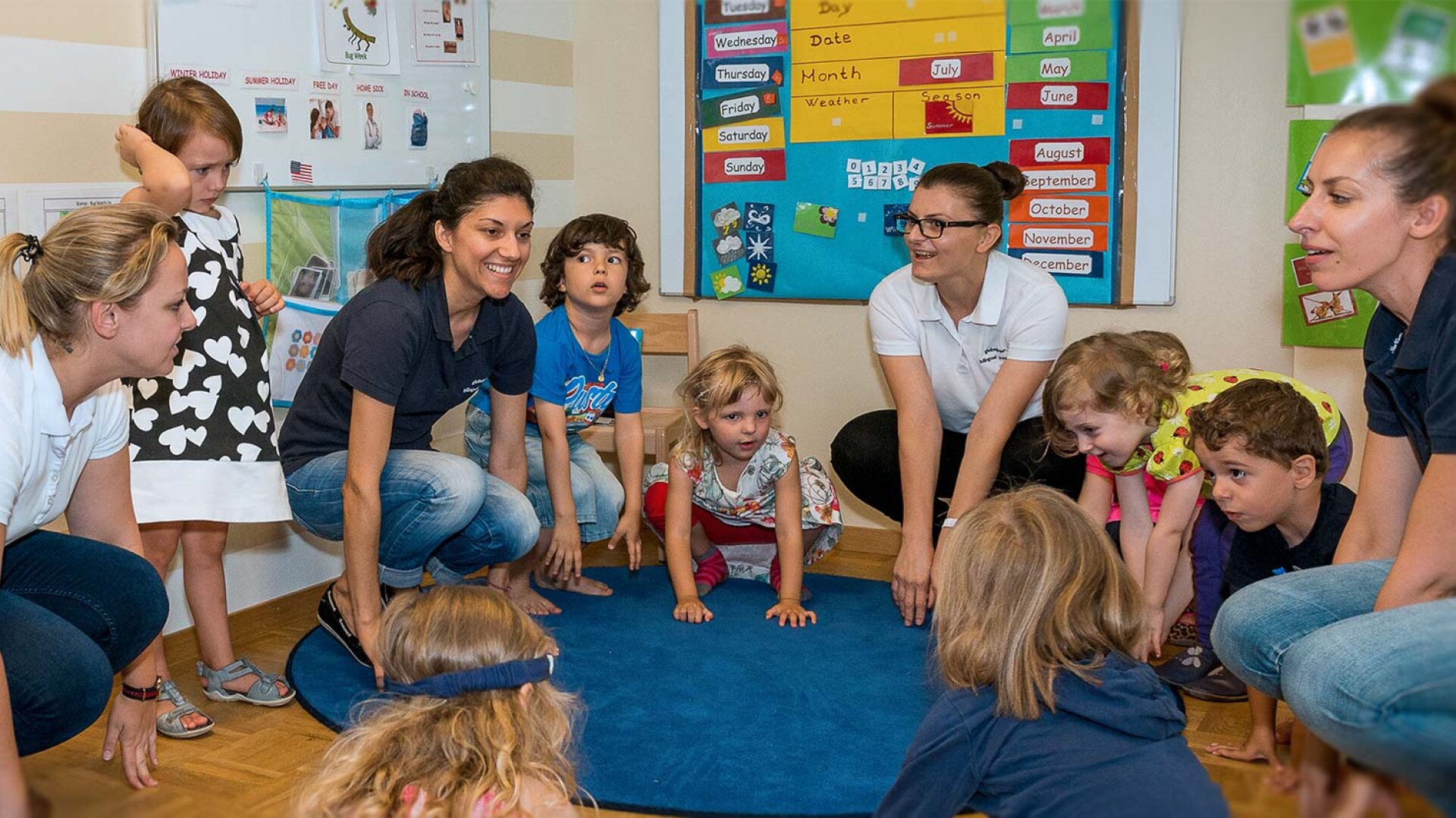 Eine Gruppe junger Kinder und vier Erwachsene sind in einem Klassenzimmer um einen blauen runden Teppich versammelt, spielen und lächeln. An den Wänden im Hintergrund hängen bunte Bildungsposter und ein Kalender.