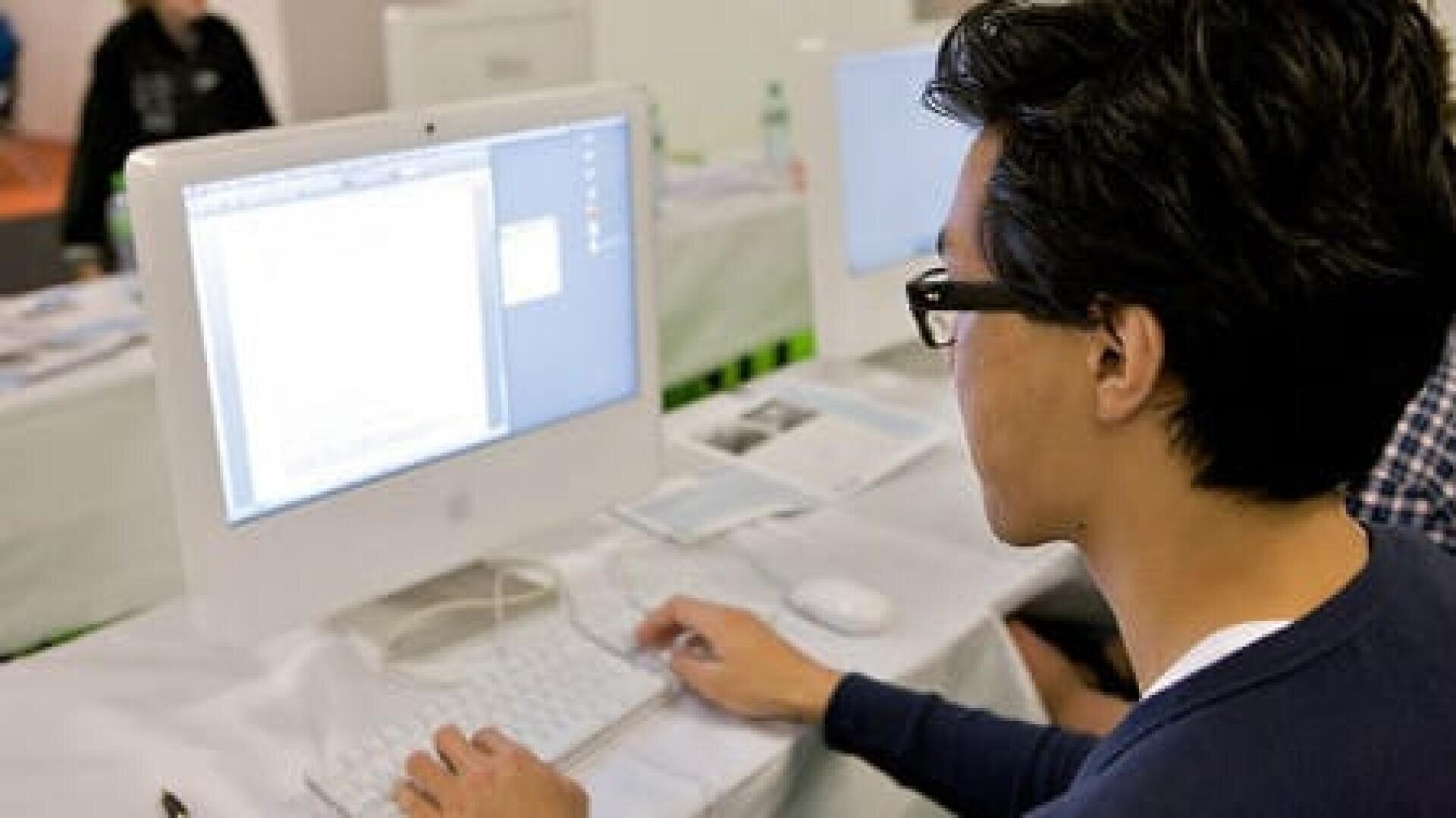 Eine Person mit dunklem Haar und Brille tippt auf einer weißen Tastatur, während sie auf einen Computermonitor schaut, auf dem ein Dokument angezeigt wird. Auf dem Tisch sind Papiere und ein weiterer Computer zu sehen.