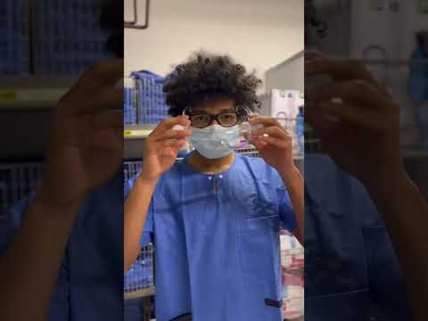 Eine Person mit lockigem Haar, die eine Brille und einen blauen Kittel trägt, legt eine chirurgische Maske in einem Raum an, der wie ein medizinischer Versorgungsraum aussieht. Im Hintergrund sind Regale mit verschiedenen Materialien zu sehen.