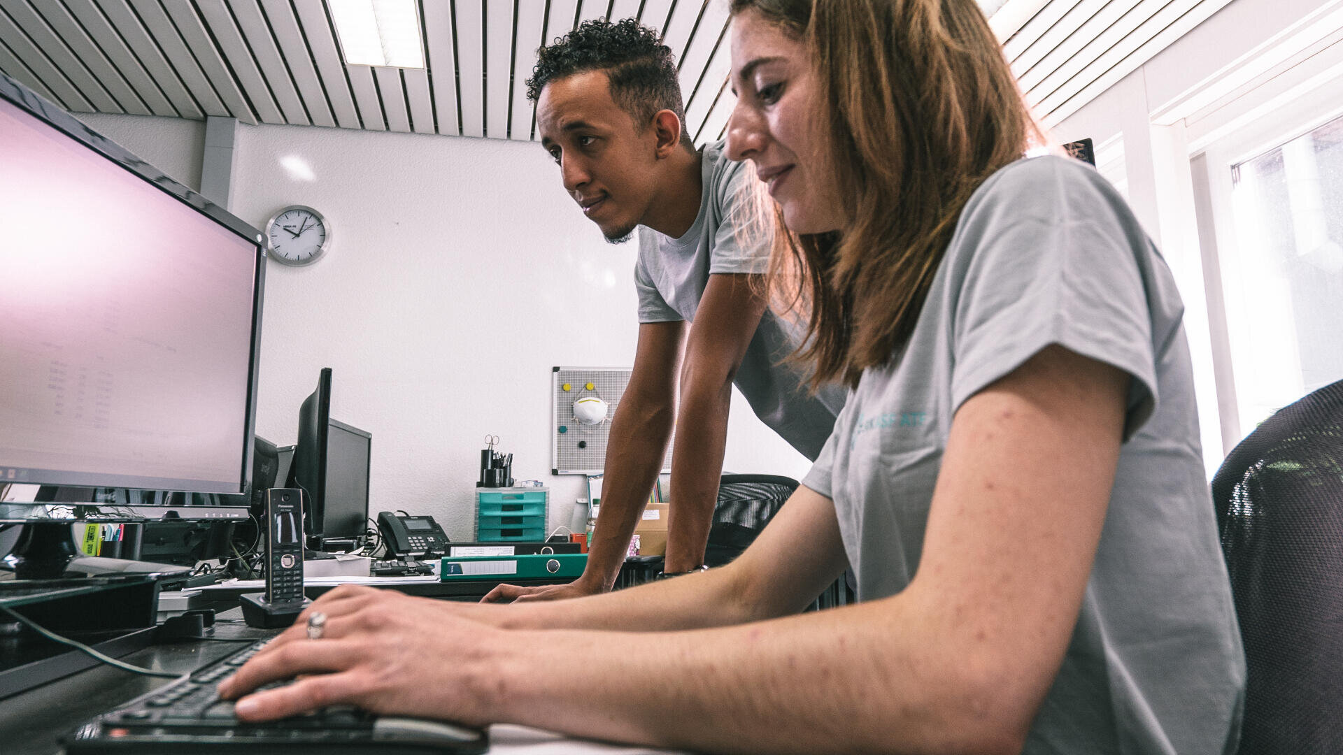 Zwei Personen arbeiten gemeinsam an einem Schreibtisch mit einem Computer. Eine Person sitzt und tippt auf der Tastatur, während die andere steht und auf den Monitor schaut. Beide tragen legere graue Hemden in einem hell erleuchteten Büro.