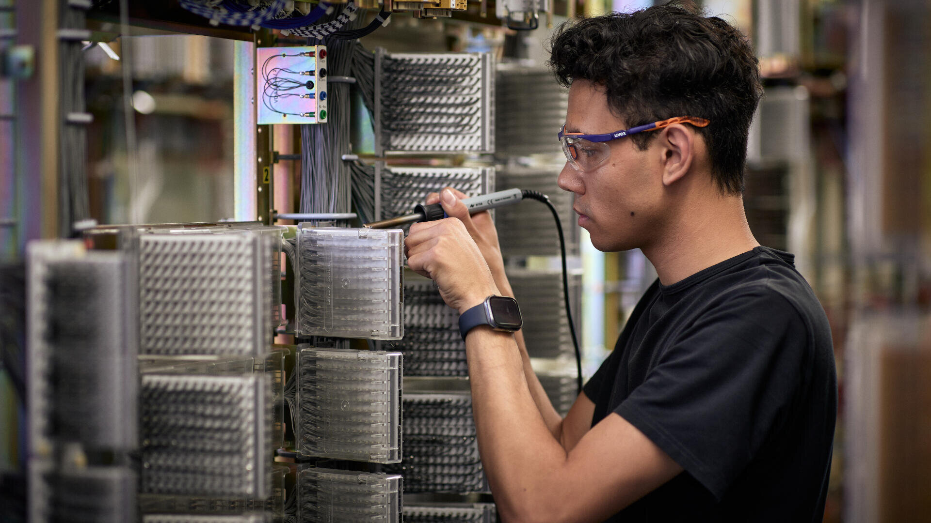 Eine Person, die eine Schutzbrille und eine Smartwatch trägt, lötet in einem Labor oder einer Werkstatt elektronische Bauteile auf einer großen Leiterplatte.