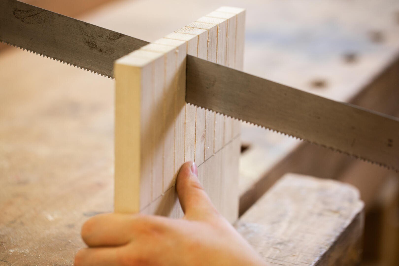 Eine Nahaufnahme einer Hand, die ein Holzstück hält, das mit einer feinzahnigen Säge entlang der gezeichneten Linien auf einer Werkbank geschnitten wird, zeigt einen Holzbearbeitungsprozess.