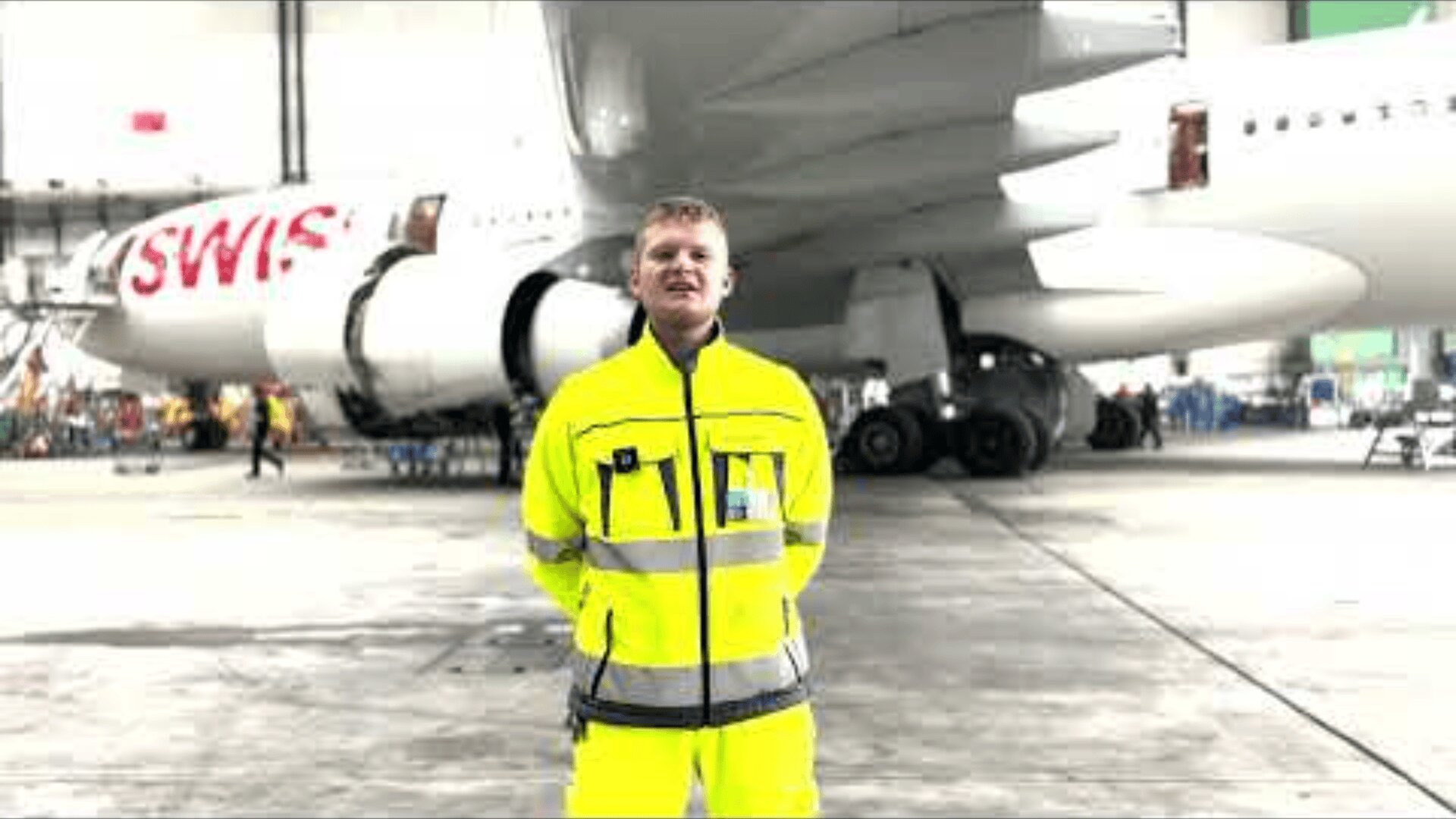 Eine Person in einem leuchtend gelben Sicherheitsanzug steht vor einem SWISS-Flugzeug in einem Flugzeughangar. Die Triebwerke und das Fahrwerk des Flugzeugs sind sichtbar, der Hangar ist geräumig und gut beleuchtet.