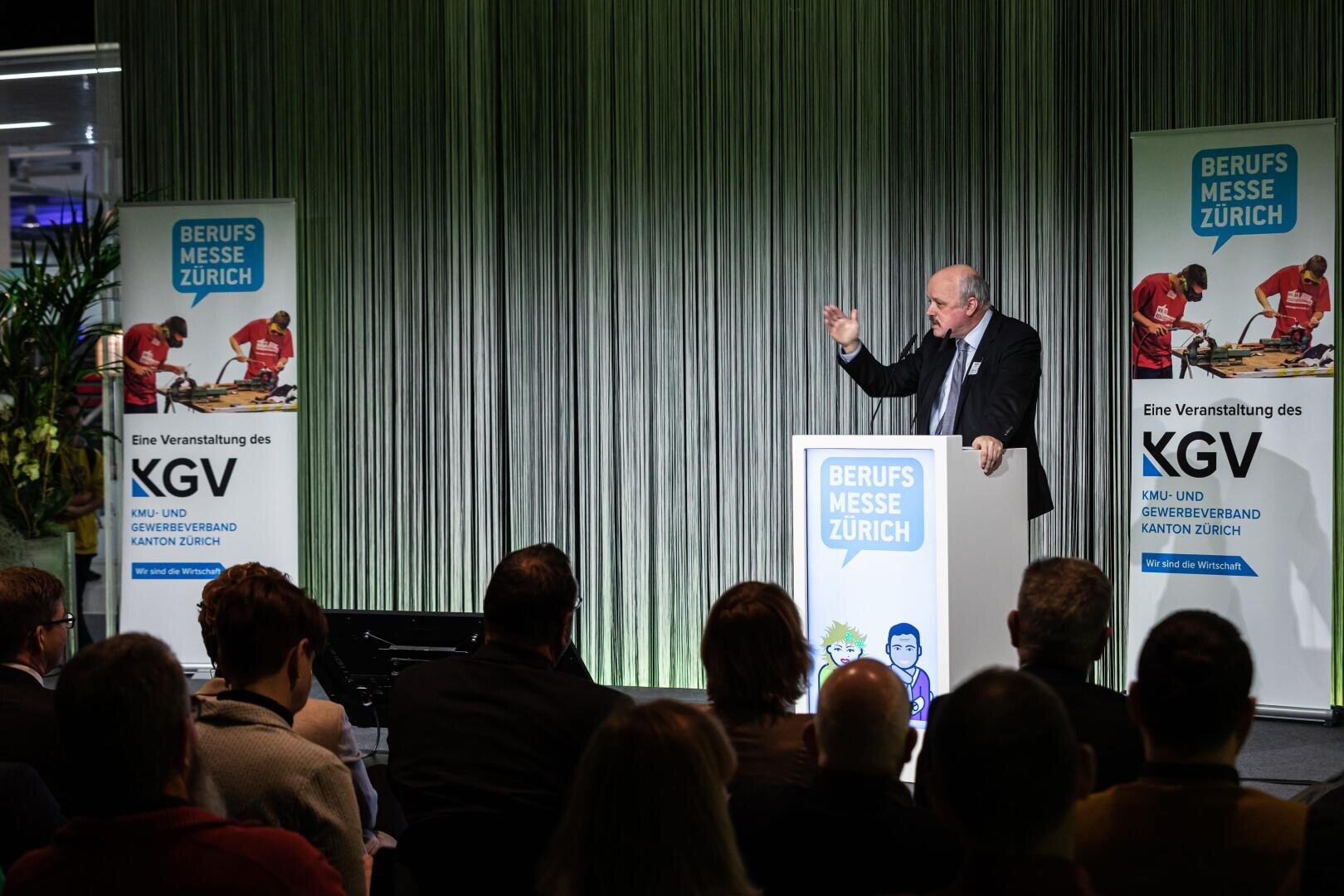 Ein Mann spricht während der Berufsmesse Zürich auf einem Podium zu den Zuhörern. Die Bühne ist mit Bannern mit dem Branding der Veranstaltung und den Logos des KGV Zürich versehen. Vertikale Vorhänge bilden den Hintergrund.
