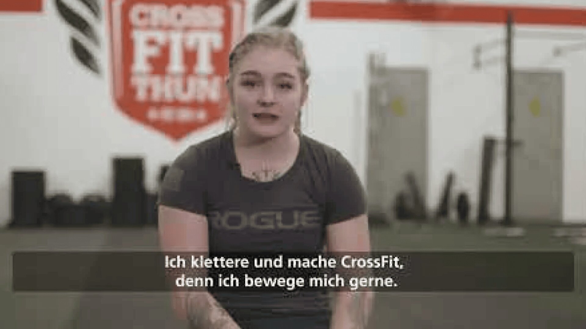 Eine junge Frau mit blondem Haar sitzt in einem Fitnessstudio und trägt ein ROGUE-T-Shirt. Im Hintergrund sind Fitnessgeräte und ein CrossFit Thun-Schild zu sehen. Die deutschen Untertitel lauten: Ich klettere und mache CrossFit, denn ich bewege mich gerne.