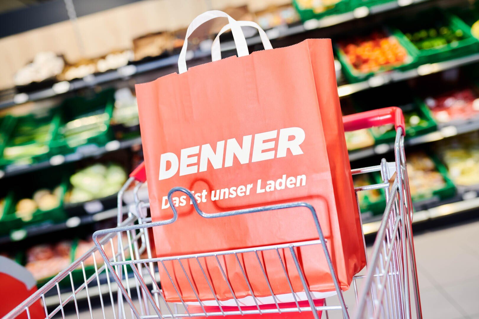 Eine rote Einkaufstasche mit dem weißen Denner-Logo und deutschem Text wird in einen Einkaufswagen vor einer Supermarktabteilung mit verschiedenen Obst- und Gemüsesorten gelegt.