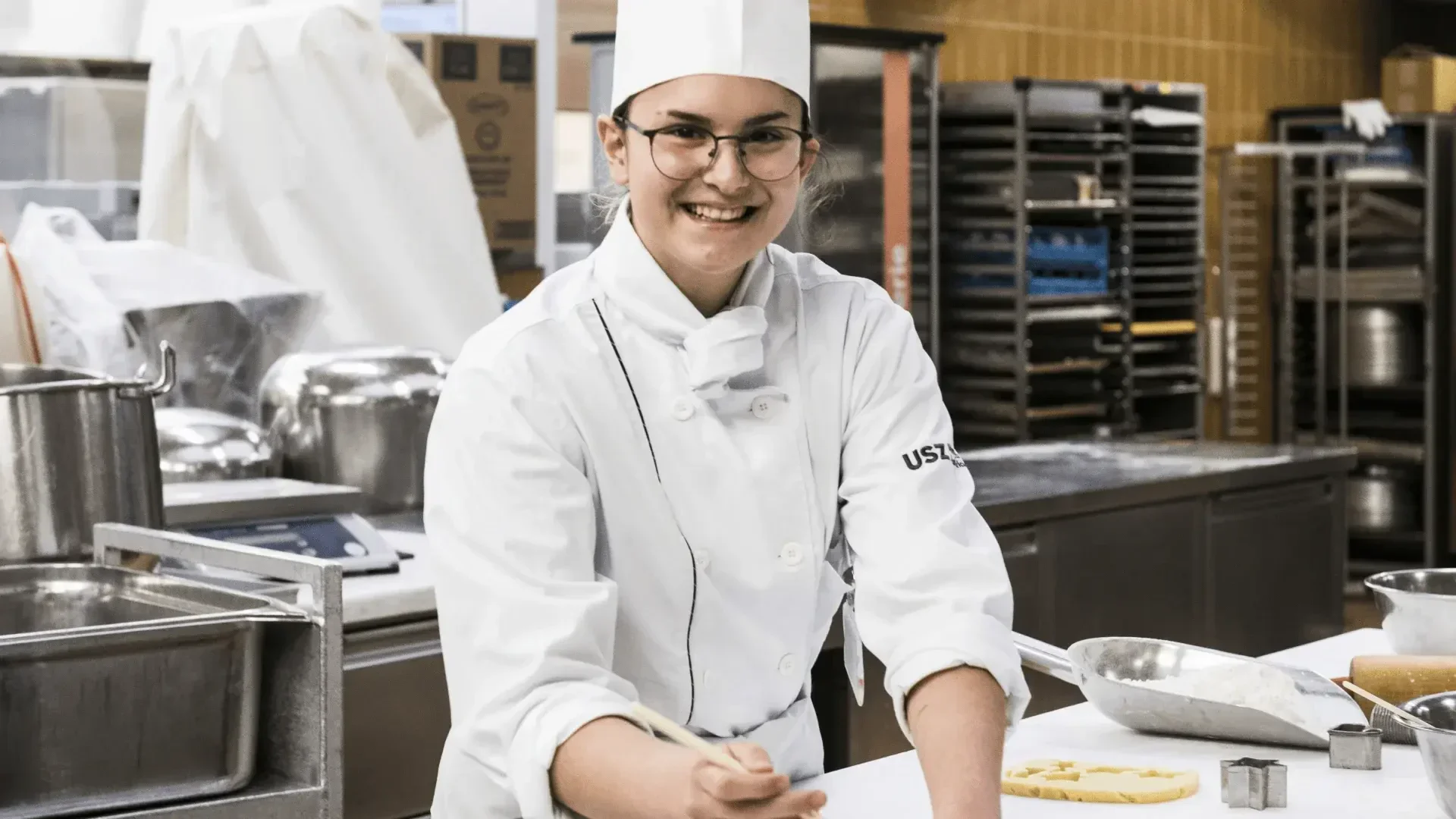 Ein lächelnder Koch in weißer Uniform und Mütze verziert Kekse in einer Großküche, umgeben von Backwerkzeugen und Zutaten.