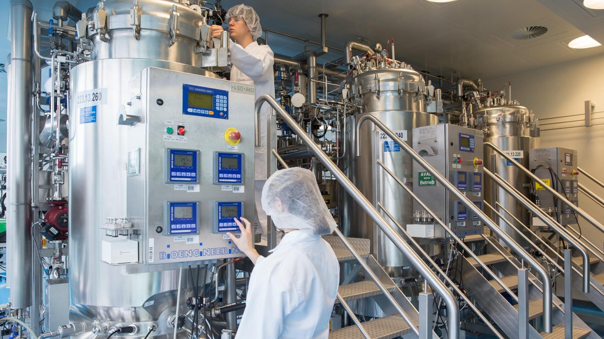 Zwei Personen in Laborkitteln und mit Haarnetzen bedienen große Bioreaktoren aus Edelstahl mit digitalen Bedienfeldern in einem sauberen, modernen Labor mit industriellen Geräten und Metalltreppen.