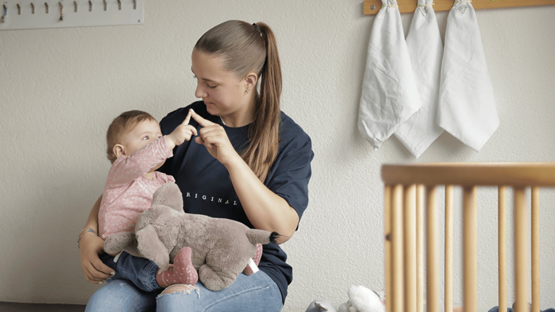Eine Frau sitzt mit einem Baby auf ihrem Schoß und berührt sanft die Zeigefinger, während das Baby einen ausgestopften Elefanten hält. Sie teilen einen zärtlichen Moment in einem Raum mit Haken und hängenden Tüchern im Hintergrund.