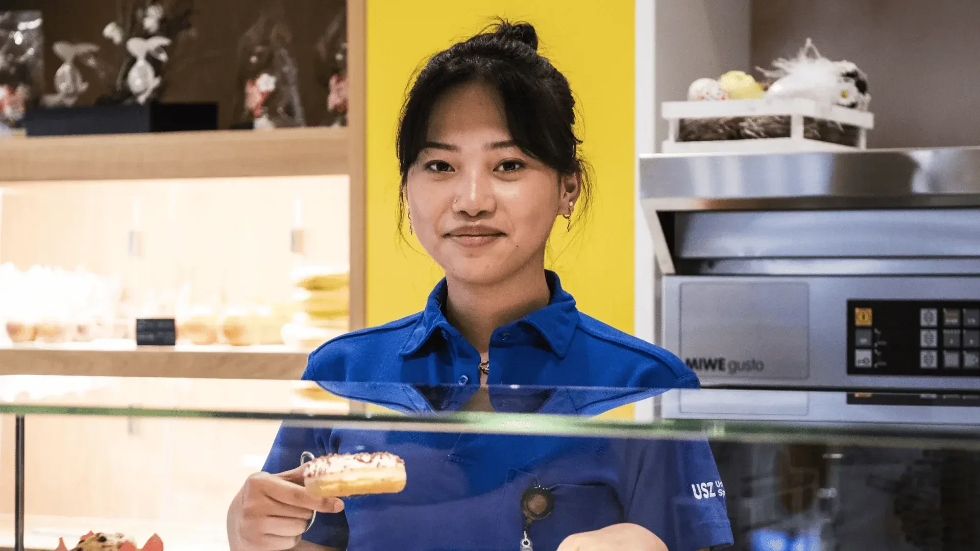 Eine Frau in blauer Uniform steht hinter einer Bäckerei, lächelt und hält ein Gebäckstück in der Hand. Im Hintergrund sind Regale mit Backwaren und ein gewerblicher Backofen zu sehen.