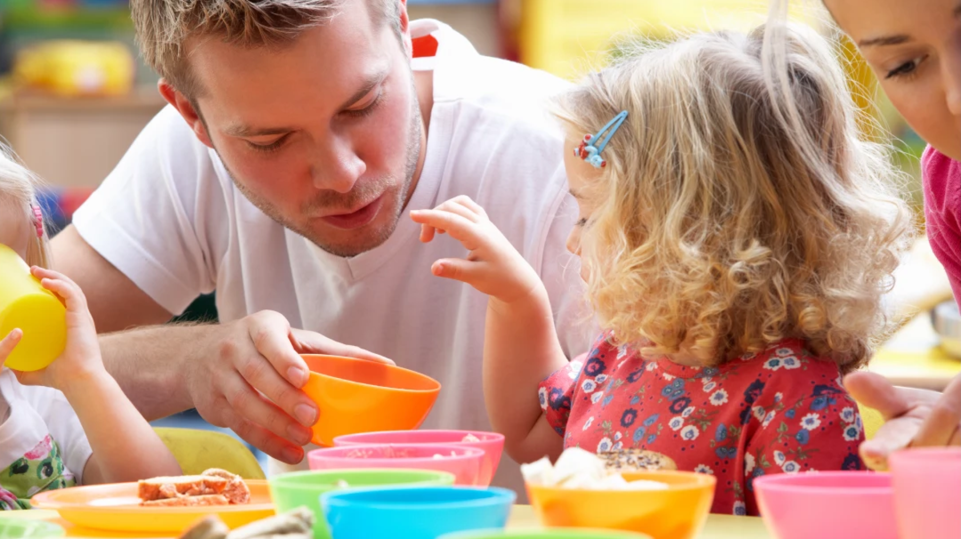 Ein Mann und ein junges Mädchen sitzen an einem Tisch mit bunten Schüsseln und teilen sich eine Mahlzeit. Der Mann spricht mit dem Mädchen, das lockiges blondes Haar hat und ein rotes Kleid mit Blumen trägt. Andere Kinder und ein Erwachsener sind teilweise zu sehen.