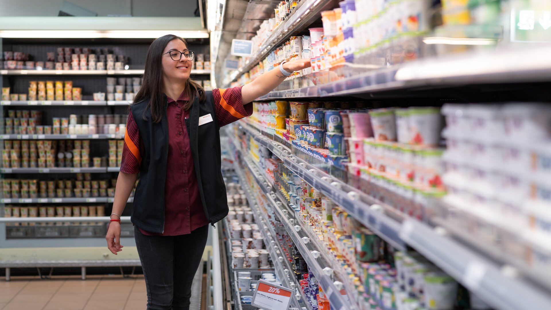 Ein Supermarktangestellter mit Brille und Namensschild lagert oder ordnet Joghurt und Milchprodukte in einem Kühlregal in einem Gang des Lebensmittelgeschäfts ein.