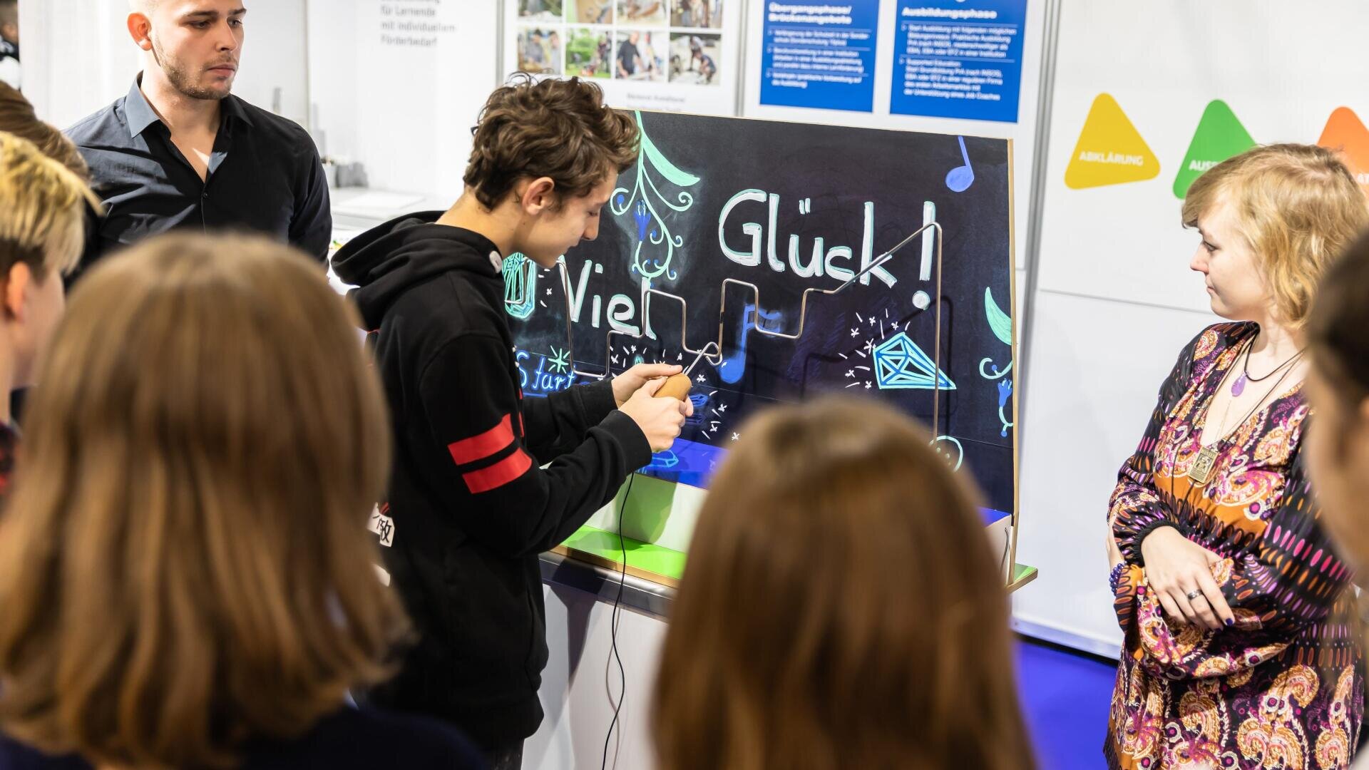 Eine Gruppe von Personen beobachtet einen jungen Mann, der ein Werkzeug an einem Schautisch mit einer dekorierten Kreidetafel mit der Aufschrift Viel Glück! und bunten Zeichnungen benutzt. Die Atmosphäre wirkt lebendig und einnehmend, möglicherweise bei einer Bildungsveranstaltung.