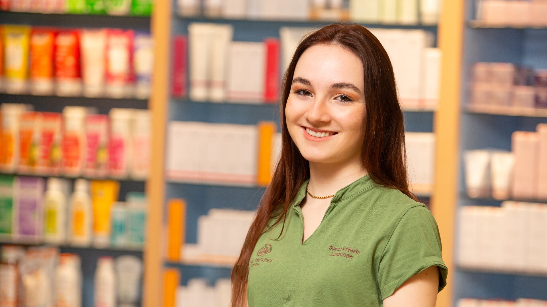 Eine junge Frau mit langen braunen Haaren und grünem Hemd lächelt, während sie in einer Apotheke oder einem Geschäft mit bunten Produkten in den Regalen im Hintergrund steht.