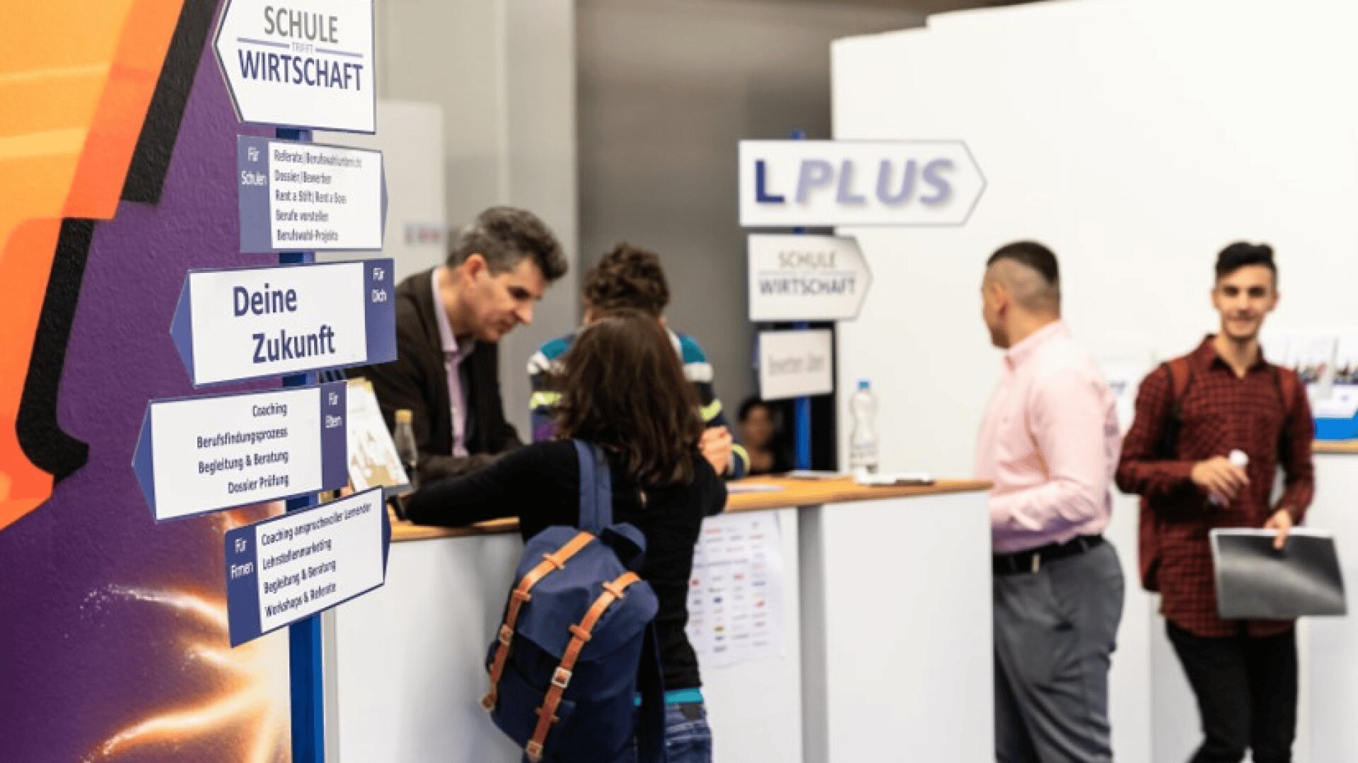 Mehrere Personen stehen an einem Informationsstand mit der Aufschrift Schule Wirtschaft und LPLUS und unterhalten sich. Auf den Schildern steht "Deine Zukunft". Eine Frau mit einem Rucksack steht vor dem Stand, und es sind Informationsflyer zu sehen.