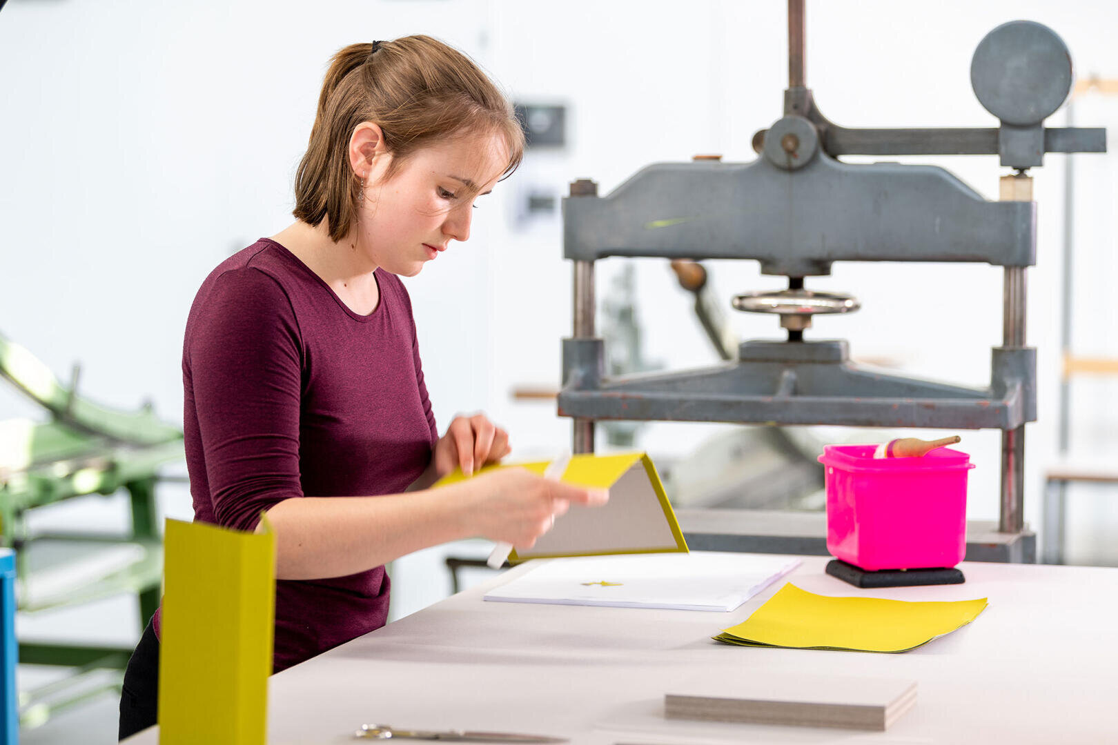 Eine Frau steht an einem Arbeitstisch und faltet gelbes Papier. Sie befindet sich in einer hellen, industriellen Umgebung mit einer großen Druck- oder Pressmaschine in der Nähe und einem rosa Behälter mit Werkzeugen auf dem Tisch.