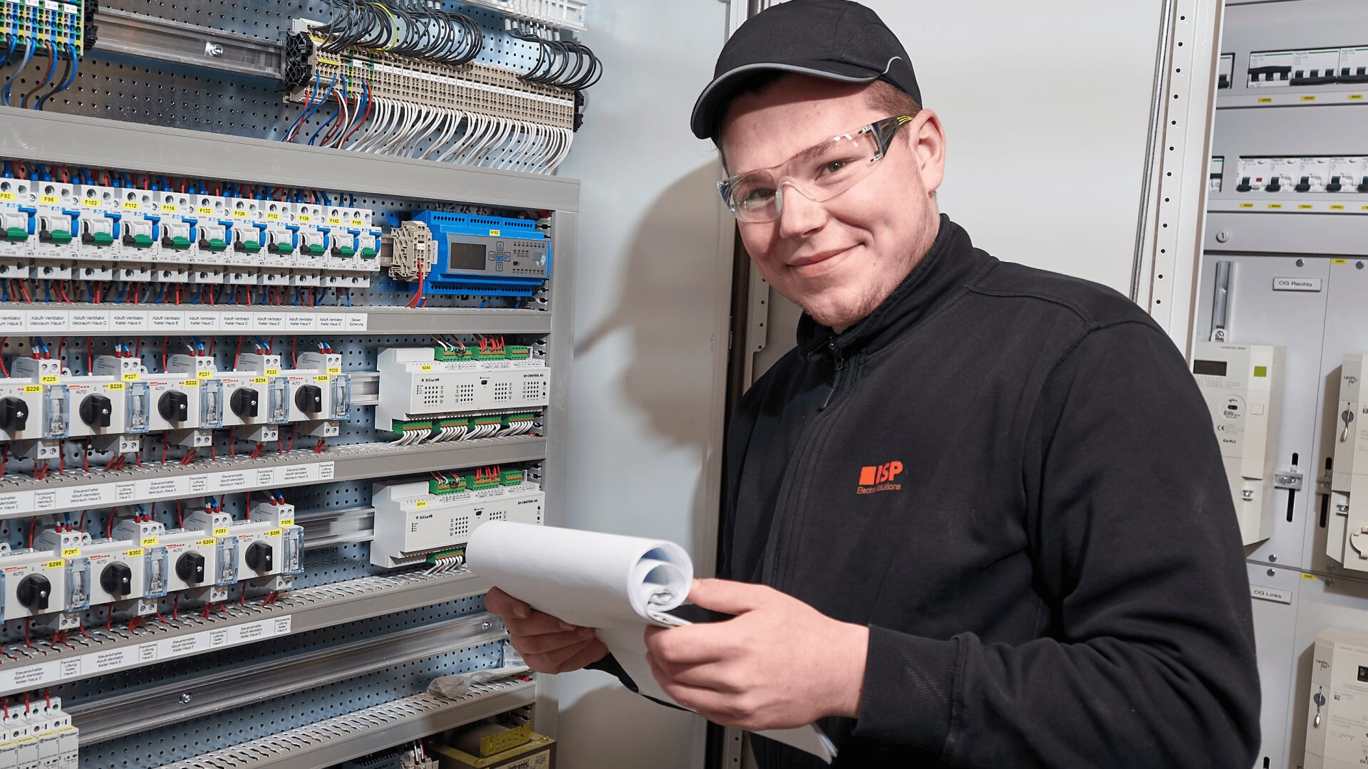 Ein Mann mit Schutzbrille, Mütze und schwarzer Jacke hält Blaupausen in der Hand und lächelt, während er neben einer elektrischen Schalttafel mit verschiedenen Schaltern und Drähten steht.