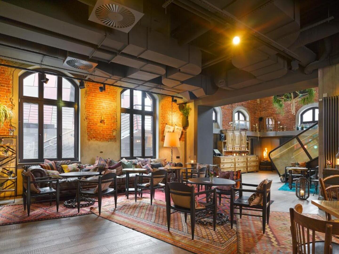 Eine gemütliche Lounge mit freiliegenden Backsteinwänden, großen Bogenfenstern, einer Mischung aus Holz- und Polstermöbeln, bunten Teppichen und Deckenrohren im Industriestil, die eine warme und einladende Atmosphäre schaffen.