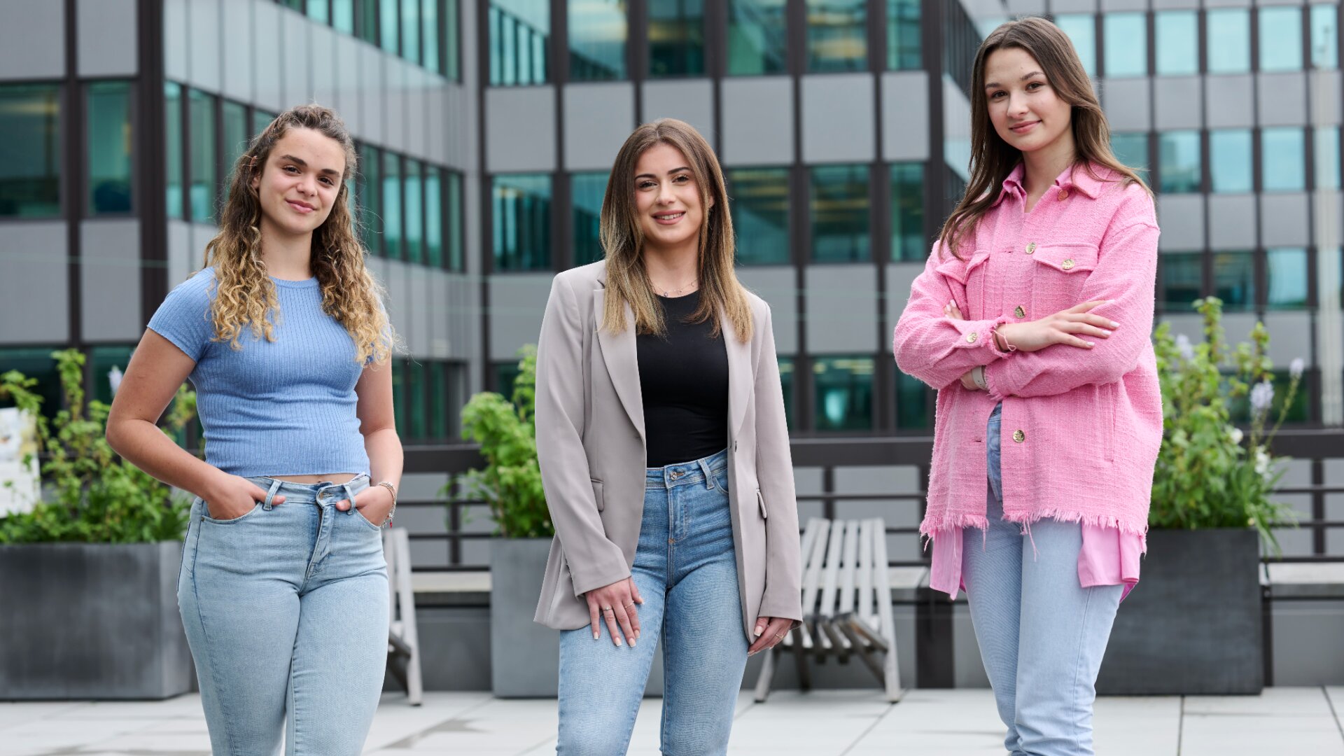 Drei junge Frauen stehen im Freien auf einem Dach, posieren und lächeln in die Kamera. Sie sind in Freizeit- und Business-Casual-Kleidung gekleidet, mit Gebäuden und Grünflächen im Hintergrund.