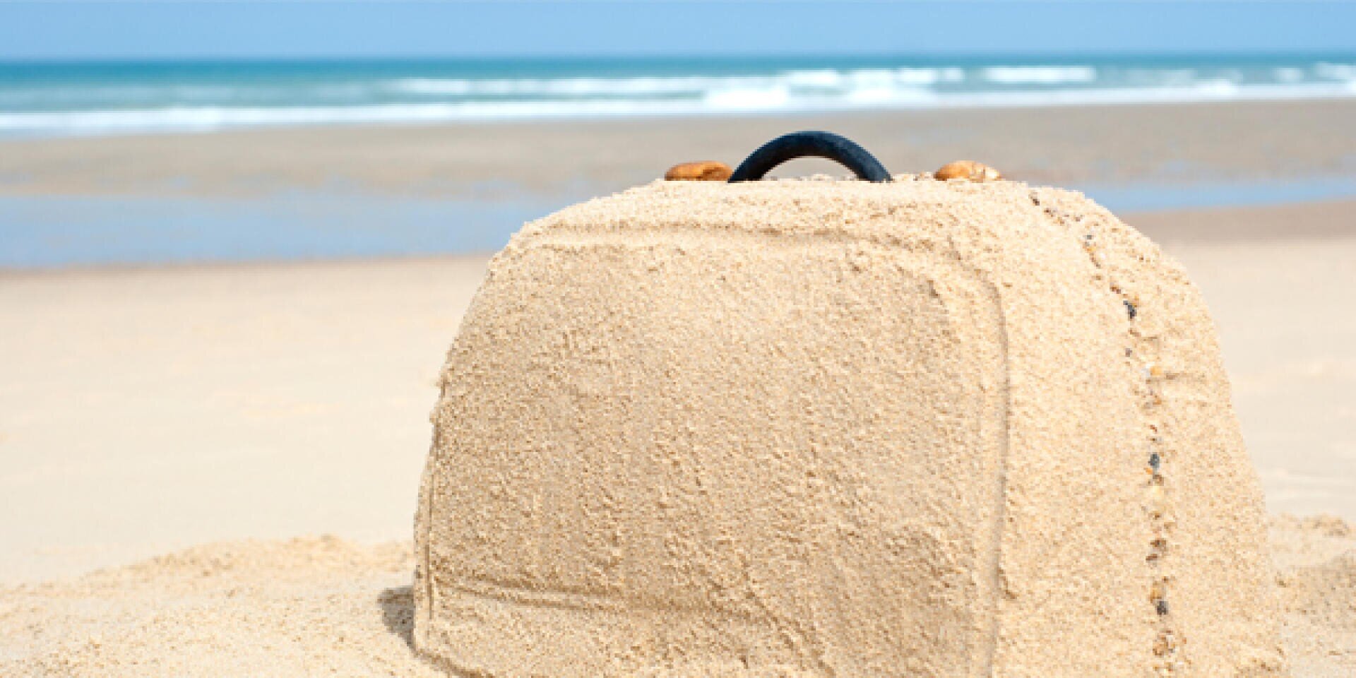 Ein mit Sand bedeckter Koffer liegt an einem Strand in der Nähe des Ozeans, während im Hintergrund die Wellen und der Himmel zu sehen sind.