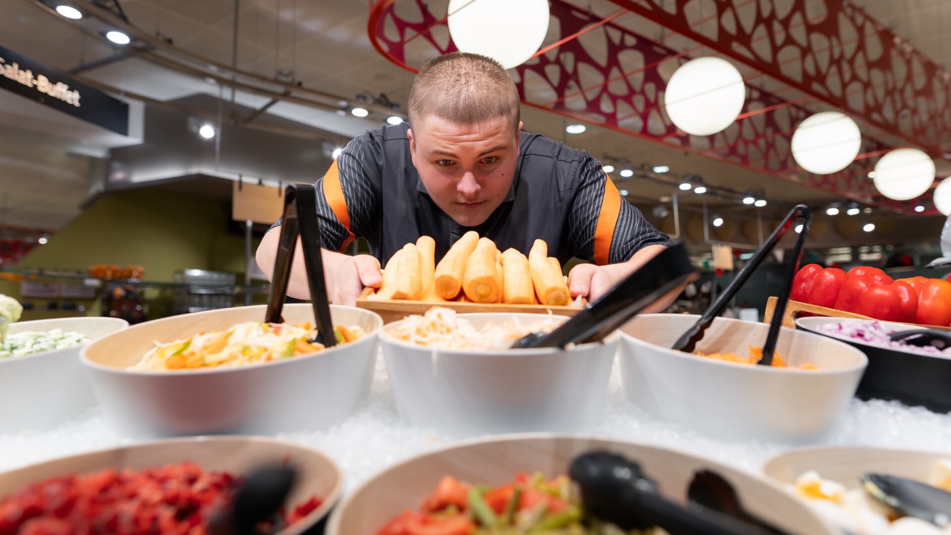 Eine Person schaut aufmerksam auf eine Salatbar und hält sich am Rand fest, während sie hinter Schüsseln mit frischem Gemüse, Belägen und Brotstangen in einer hell erleuchteten Buffetumgebung steht.