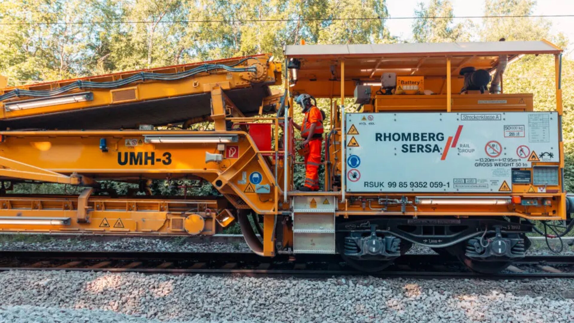 Ein Arbeiter in orangefarbener Uniform bedient eine große gelbe Eisenbahnwartungsmaschine mit der Aufschrift UMH-3 und Rhomberg Sersa auf Bahngleisen, umgeben von Bäumen und Sonnenlicht.