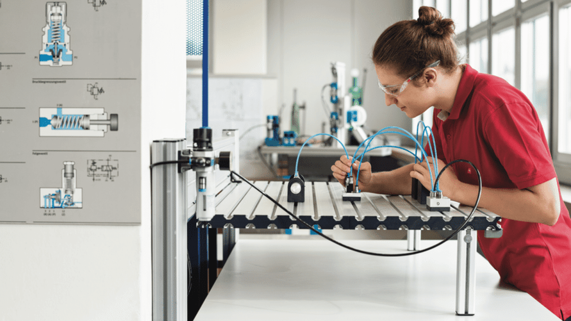 Eine Person, die eine Schutzbrille trägt, arbeitet an einem Labortisch mit pneumatischen Komponenten und passt Kabel oder Schläuche an. An einer Wand in der Nähe sind technische Diagramme angebracht, und der Raum ist durch große Fenster gut beleuchtet.