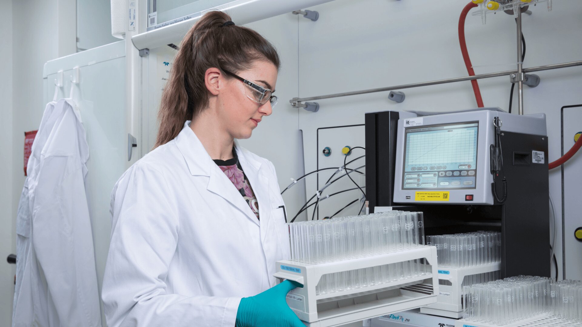 Eine Frau im Laborkittel und mit Schutzbrille hält in einem Labor ein Tablett mit Reagenzgläsern und steht vor einem wissenschaftlichen Gerät mit Digitalanzeige und aufgewickelten Kabeln.