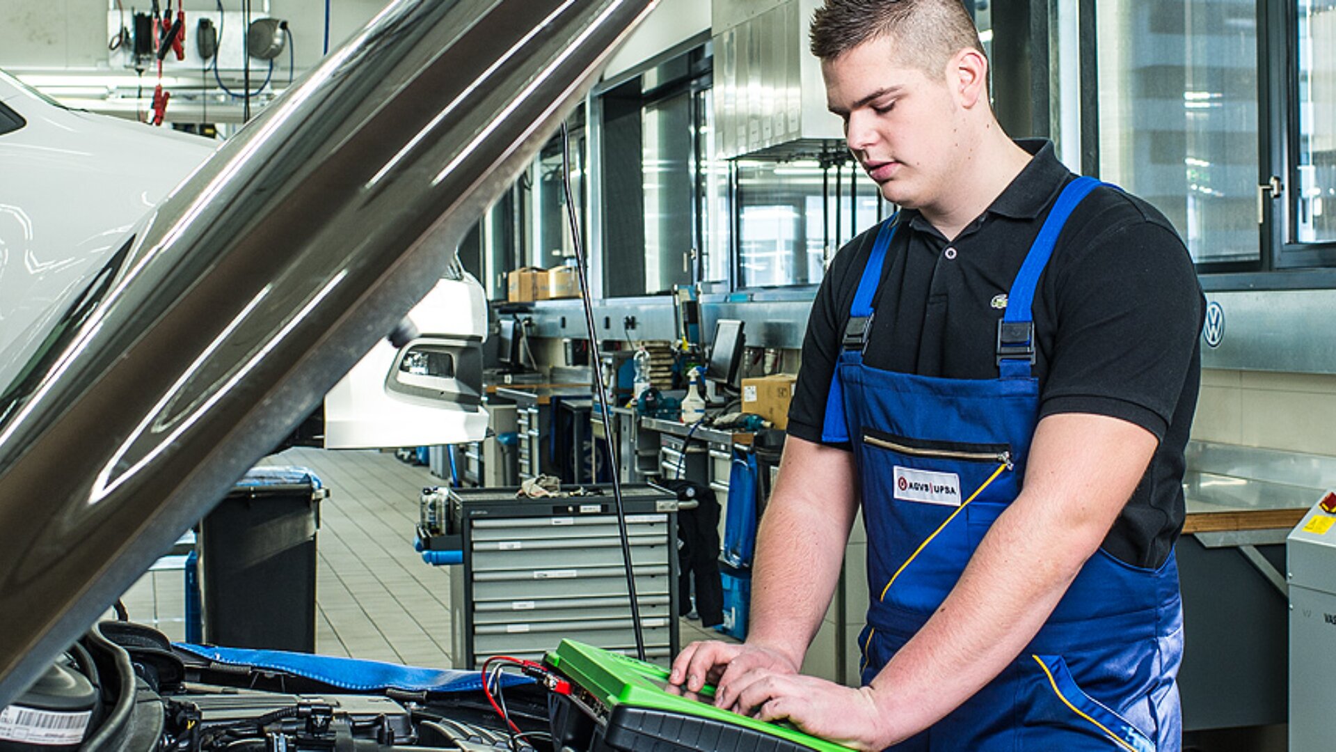 Ein Mechaniker in einem blauen Overall arbeitet mit einem Diagnosewerkzeug an einem Automotor in einer gut beleuchteten Autowerkstatt, wobei im Hintergrund verschiedene Werkzeuge und Geräte zu sehen sind.