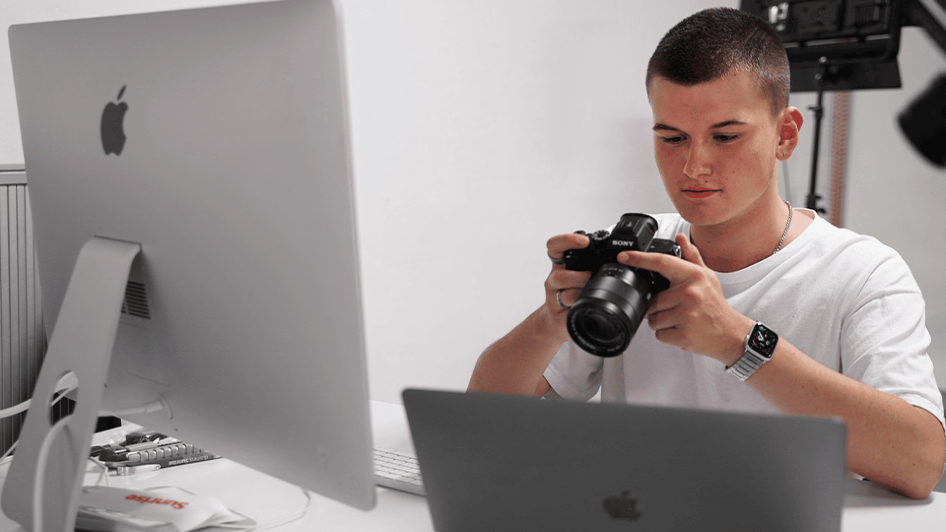Ein junger Mann in einem weißen Hemd sitzt an einem Schreibtisch mit einem iMac und einem MacBook und untersucht eine Sony-Kamera. Auf dem Schreibtisch sind Büromaterialien zu sehen, und im Hintergrund befindet sich eine Fotoausrüstung.