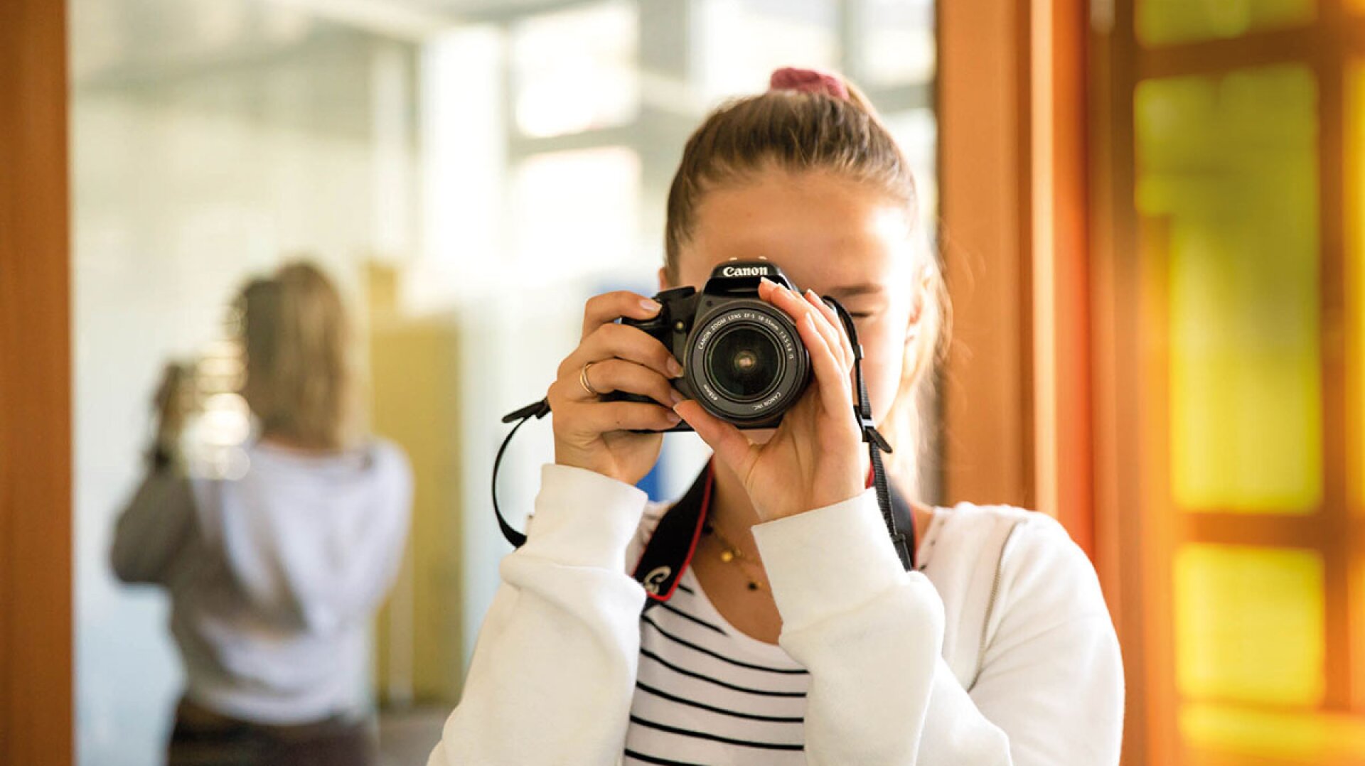Eine Person in einer weißen Jacke und einem gestreiften Hemd hält eine Canon-Kamera an ihr Gesicht und macht ein Foto in einem Innenraum. Im Hintergrund ist ein verschwommenes Spiegelbild der Person zu sehen.
