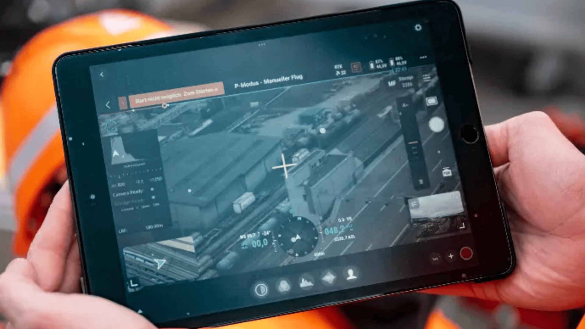 Eine Person hält ein Tablet in der Hand, auf dem eine Live-Luftaufnahme von Industriegebäuden und Fahrzeugen zu sehen ist, die wahrscheinlich von einer Drohne aufgenommen wurde, wobei Flugdaten und Steuerungen auf dem Bildschirm sichtbar sind.
