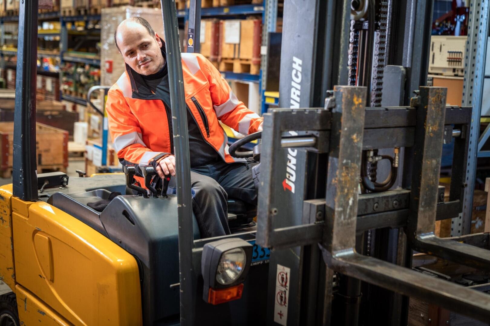 Ein Mann mit einer orangefarbenen Sicherheitsjacke bedient einen gelben Gabelstapler in einem Lager, das mit Regalen und verschiedenen Kisten gefüllt ist.