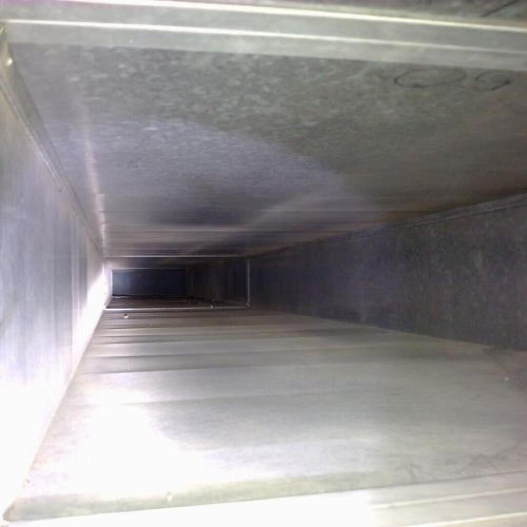 Blick in einen sauberen, rechteckigen Luftkanal aus Metall, an dessen hinterem Ende Licht zu sehen ist. Die glatten, glänzenden Oberflächen des Kanals reflektieren etwas Licht, und es sind weder Staub noch Ablagerungen sichtbar.